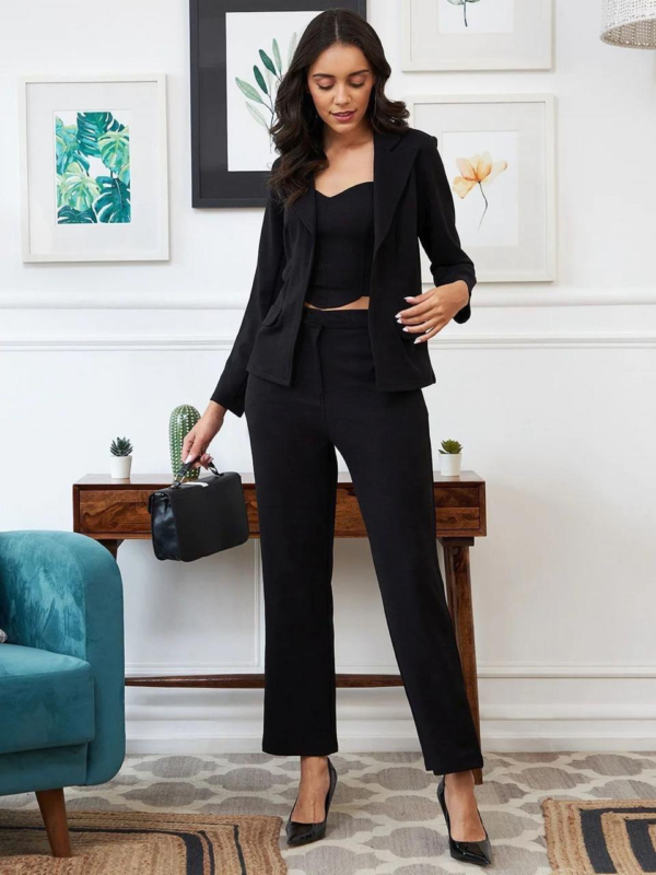 black three-piece suit featuring a tailored blazer, a trendy crop top, and straight-fit pants, paired with black heels and a handbag in a modern indoor setting