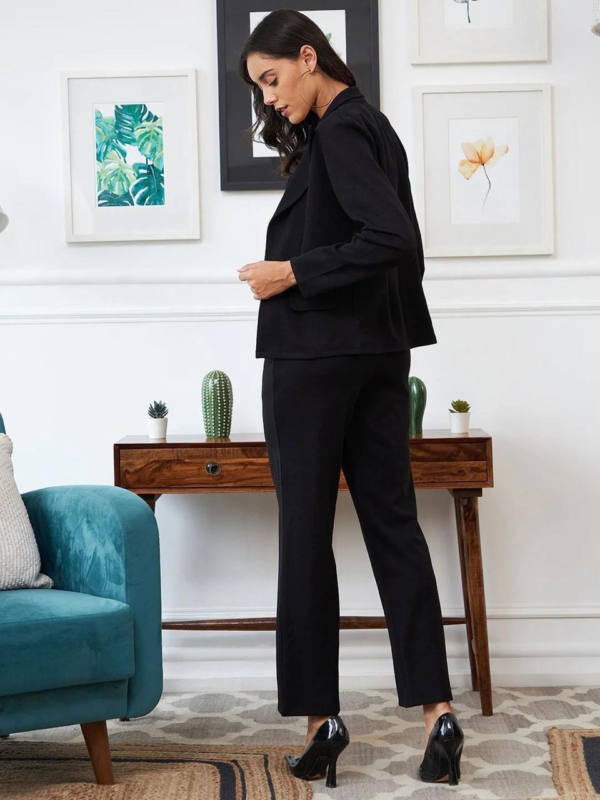 black three-piece suit featuring a tailored blazer, a trendy crop top, and straight-fit pants, paired with black heels and a handbag in a modern indoor setting