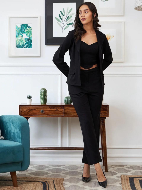 black three-piece suit featuring a tailored blazer, a trendy crop top, and straight-fit pants, paired with black heels and a handbag in a modern indoor setting