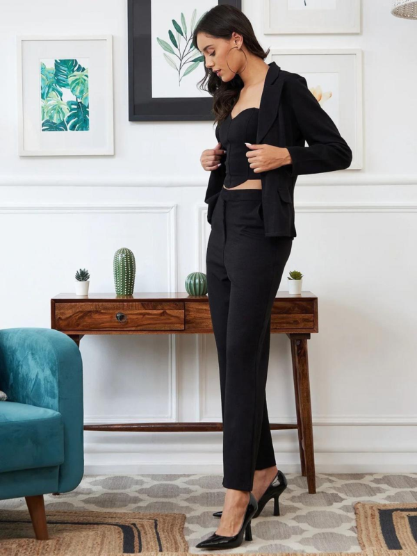 black three-piece suit featuring a tailored blazer, a trendy crop top, and straight-fit pants, paired with black heels and a handbag in a modern indoor setting