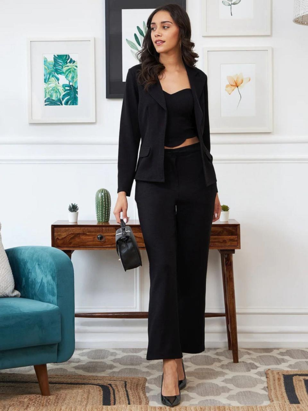 black three-piece suit featuring a tailored blazer, a trendy crop top, and straight-fit pants, paired with black heels and a handbag in a modern indoor setting